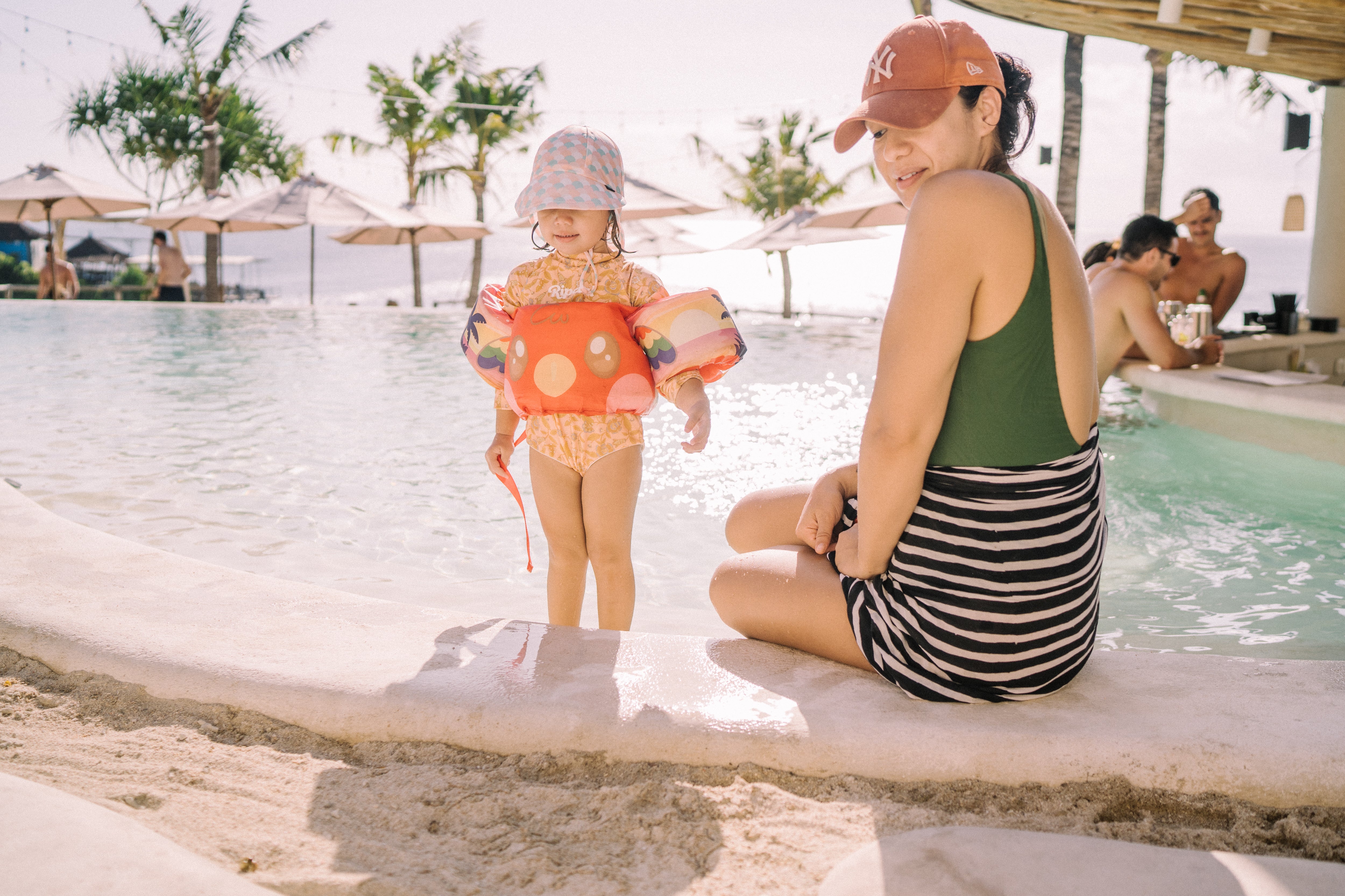 A child in a colorful floatie and hat stands by a pool, while an adult in a swimsuit and cap sits nearby.