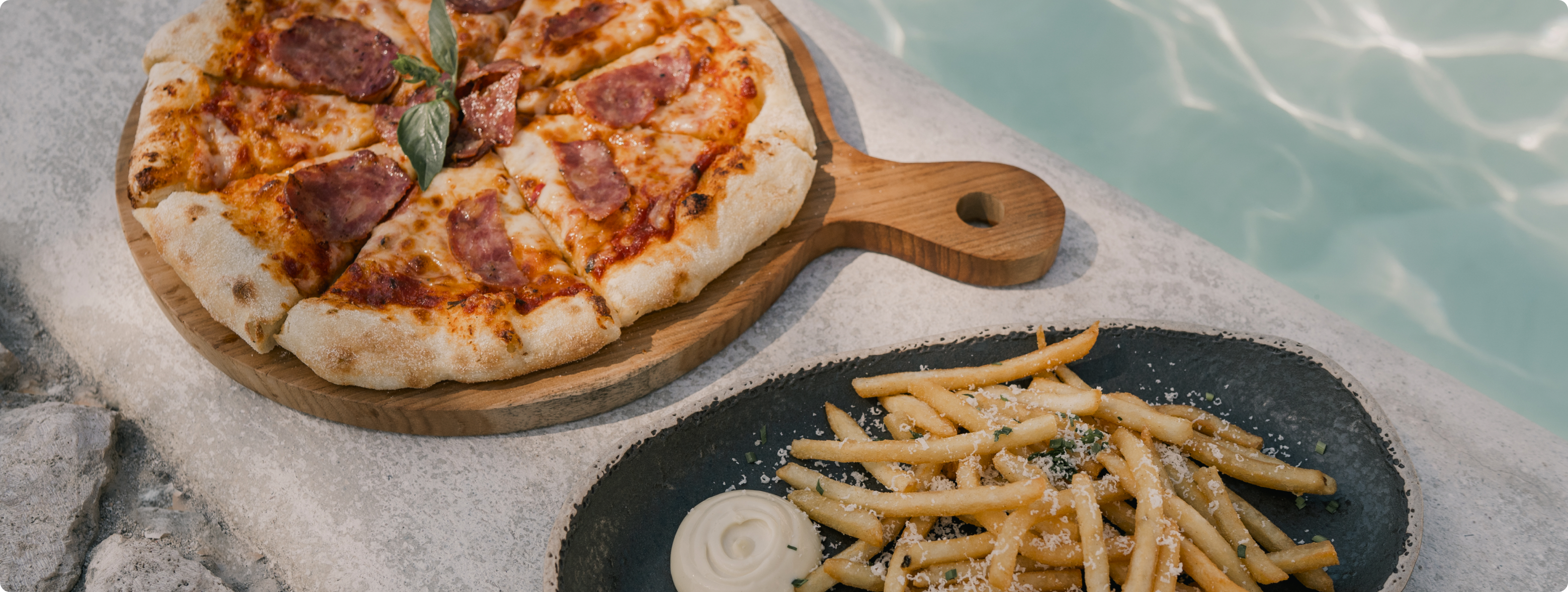 A delicious pizza and crispy French fries placed on a table beside a sparkling pool.