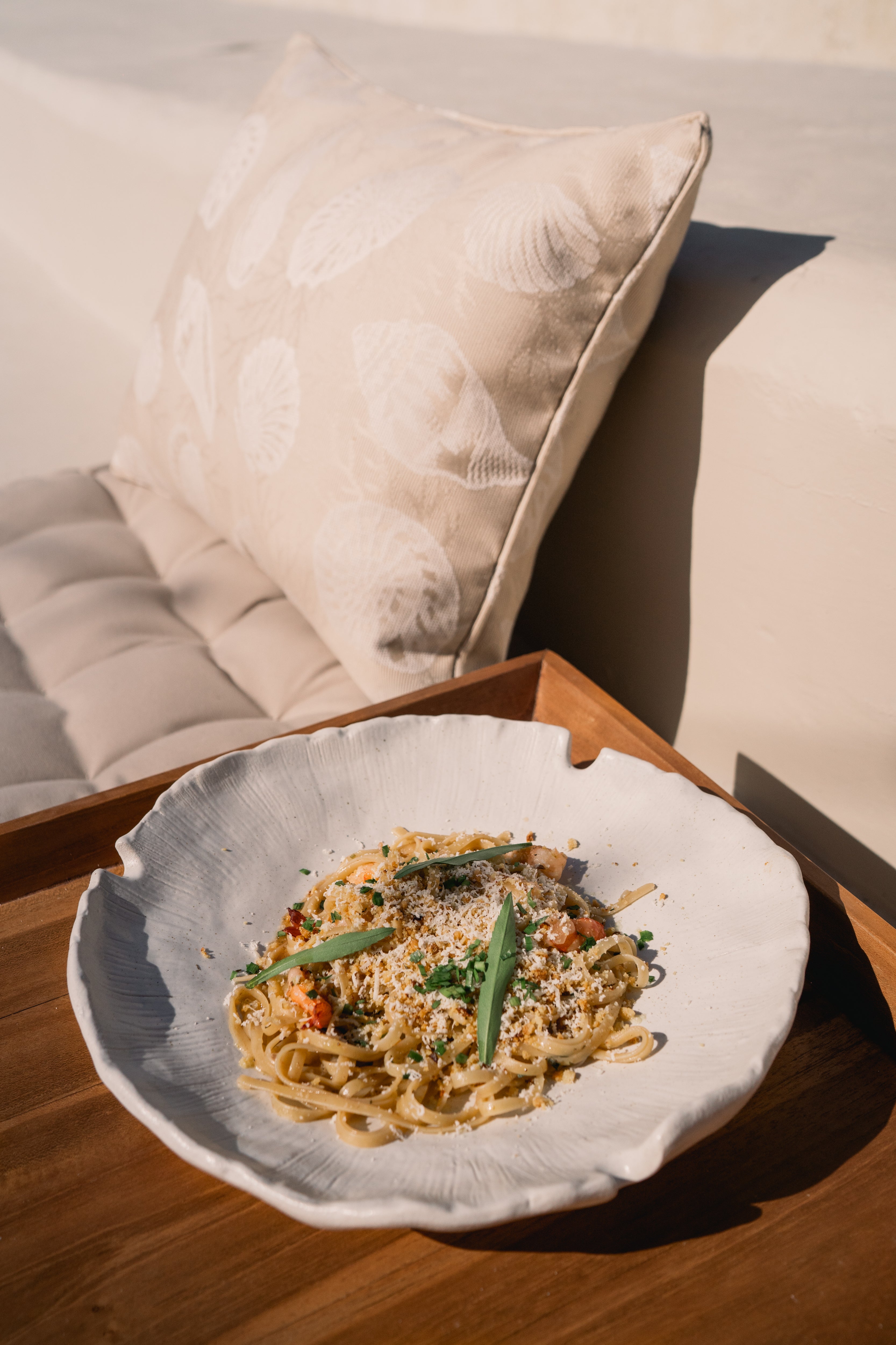 Pasta dish with herbs and cheese on a wooden tray, next to a shell-patterned cushion. 